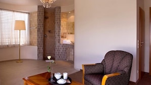 Une chambre d’hôtel dotée d’un coin salon comprenant un fauteuil à motifs, une table basse en bois sur laquelle se trouve un plateau avec des tasses blanches et un vase contenant une seule fleur, ainsi qu’un lustre.