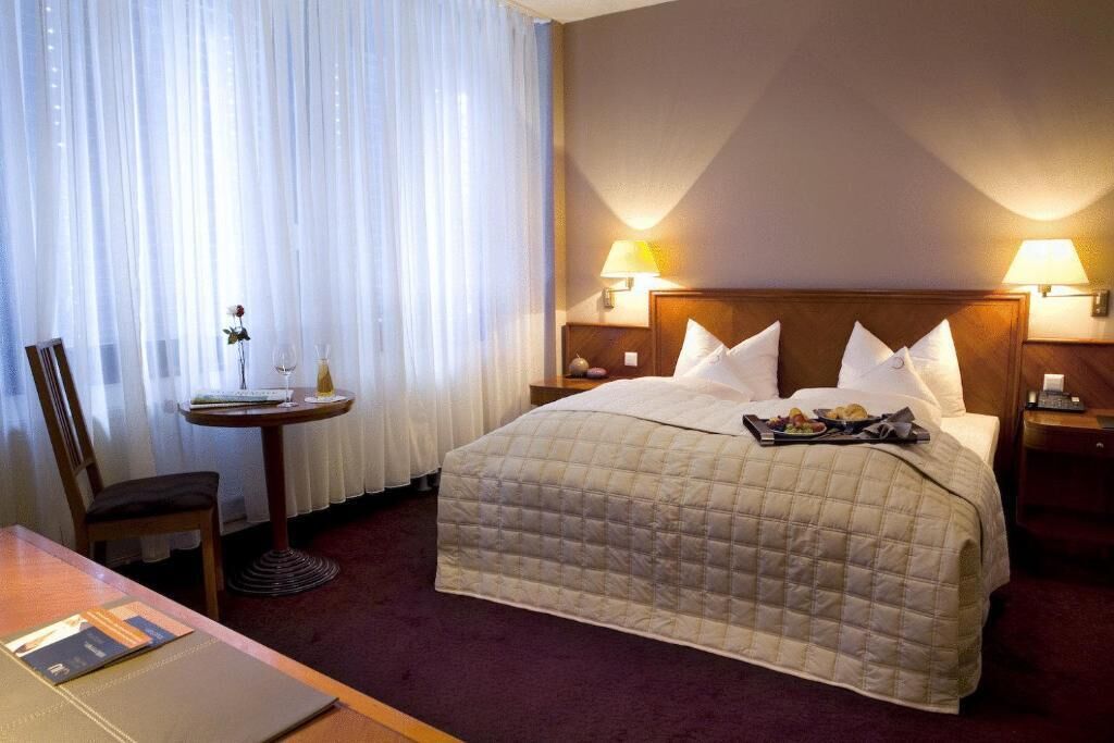 Hotellihuoneessa on sänky, kaksi yöpöytää, joissa on lamput, pieni pöytä, jossa on maljakko ja viinilaseja, sekä tuoli.