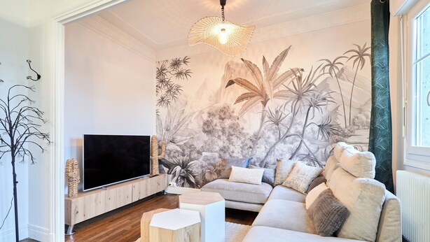 Ein modernes Wohnzimmer mit Flachbildfernseher, einer Couch, einem Couchtisch und einer Wandmalerei mit tropischem Grün.