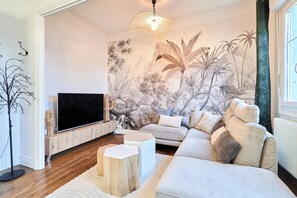 Ein modernes Wohnzimmer mit Flachbildfernseher, einer Couch, einem Couchtisch und einer Wandmalerei mit tropischem Grün.