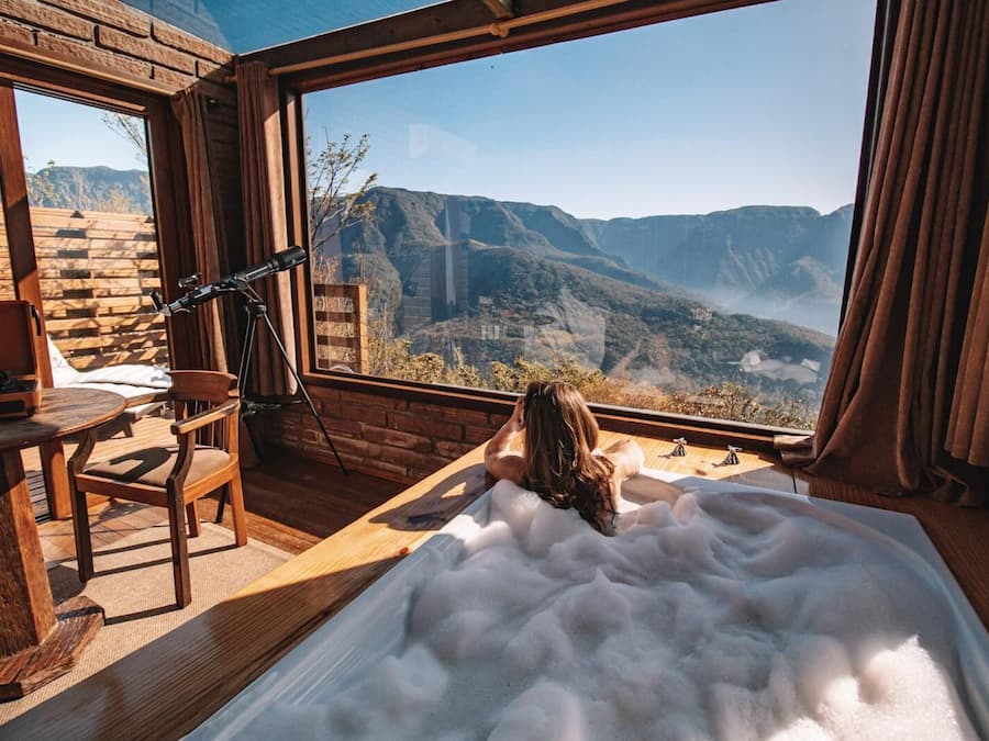 Una stanza con una grande finestra che si affaccia su un paesaggio montuoso, una scrivania in legno con un portatile e una vasca da bagno piena di bolle.