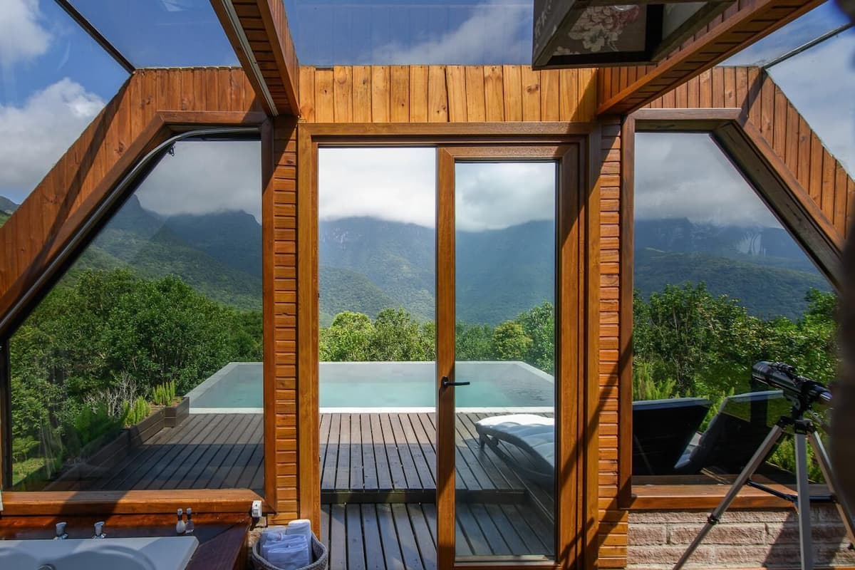Eine Holzdiele mit Glasumrandung mit Blick auf eine gebirgige Landschaft und ein Infinity-Pool.