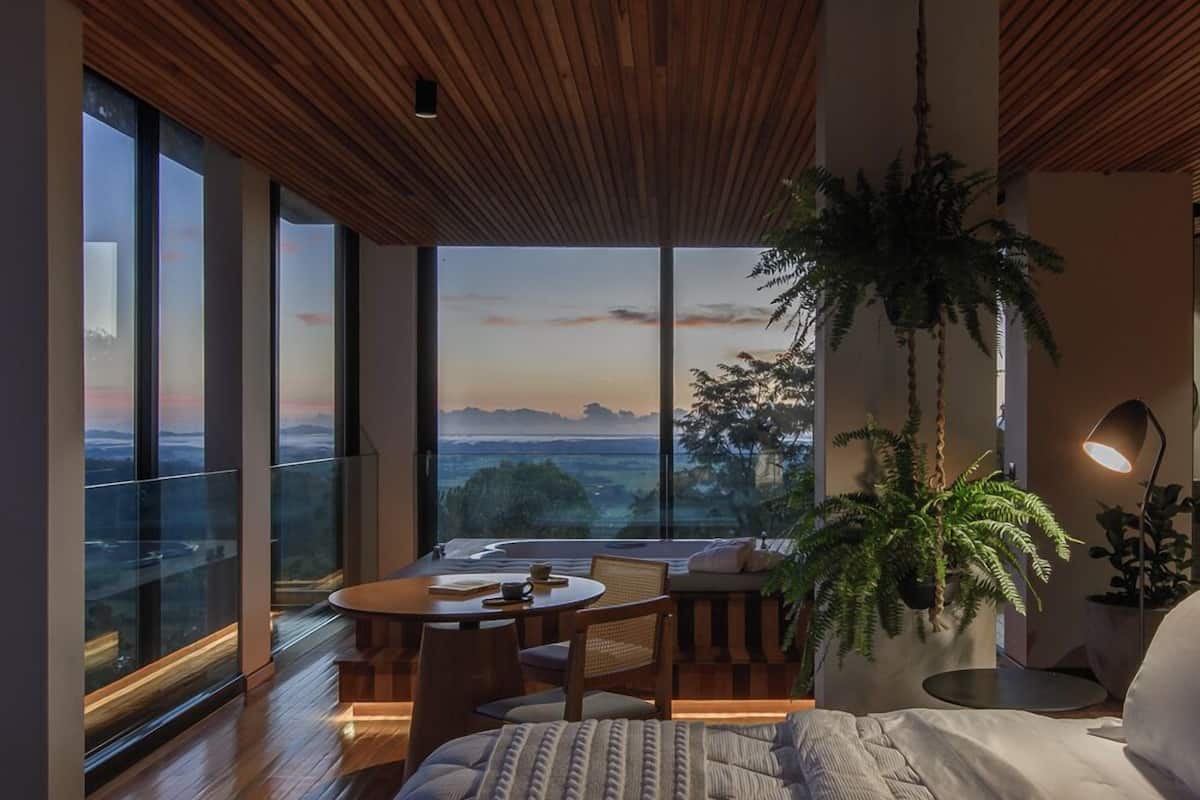 Ein modernes Schlafzimmer mit einem großen Bett, einem Esstisch und Blick auf die Landschaft durch große Fenster.