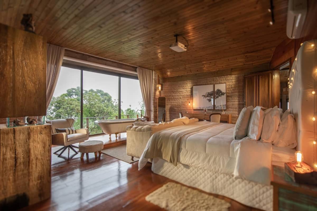 Ein geräumiges Schlafzimmer mit einem großen Bett, einer Sitzecke mit Sessel und kleinem Tisch, einer Badewanne und einem großen Fenster mit Blick auf Bäume.