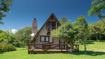 Una cabaña pequeña de dos pisos con un balcón de madera y una chimenea, rodeada de abundante vegetación.