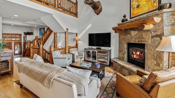 Een ruime woonkamer met een stenen openhaard, een opgehangen elandshoofd, een houten trap en een grote flatscreentelevisie.