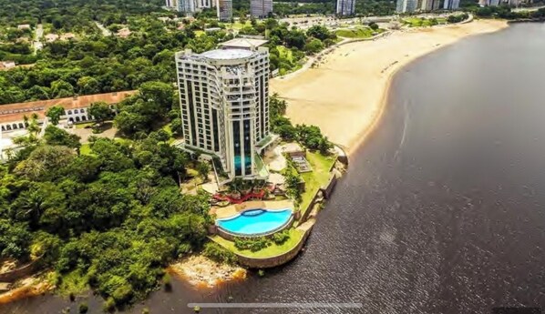 An aerial view of a coastal hotel with a swimming pool, beachfront, and lush greenery.