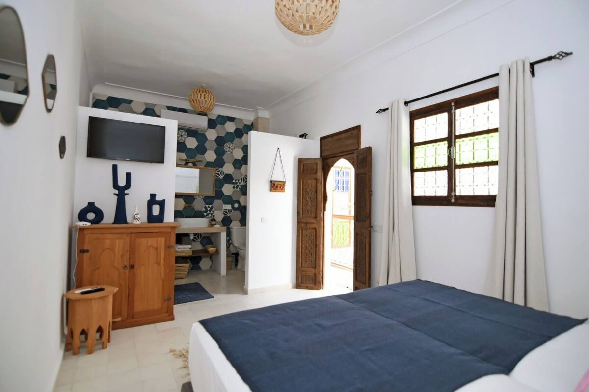 A bedroom with a bed, a TV, a wooden cabinet, and a bathroom visible through an open door.