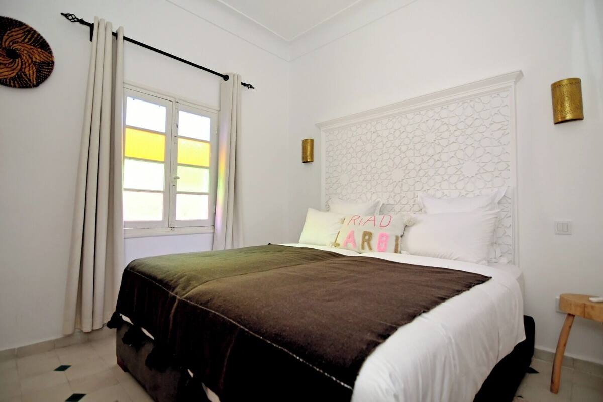 A bedroom with a bed, a window with curtains, a headboard with a geometric pattern, and a small bedside table.