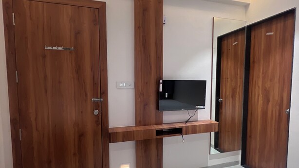 A modern bedroom with wooden wardrobes, a flat-screen TV mounted on a wooden shelf, and a bed with white bedding. - HOTEL RUDRAKSH (Rahata)