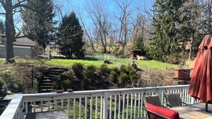 A wooden deck with a table and red chairs, overlooking a garden with a lawn, trees, and a stone structure.