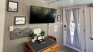 A room with a flat-screen TV mounted on the wall, a wooden desk with a chair, a small table with potted plants, and a door with curtains.