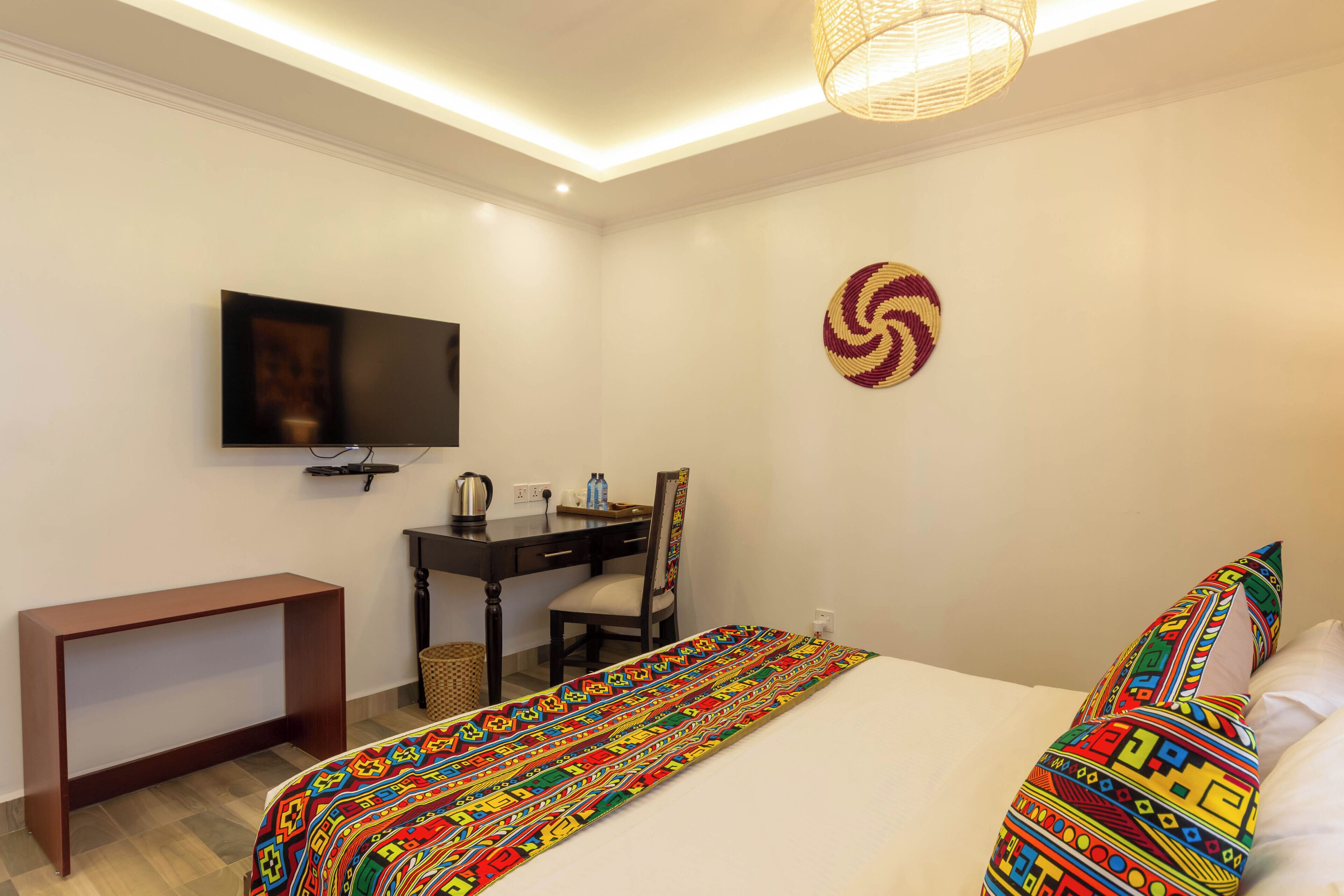 A hotel room with a bed, a desk, a chair, a television, and a woven light fixture.