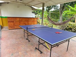 Una mesa de ping pong al aire libre con un banco de madera y una hamaca al fondo.