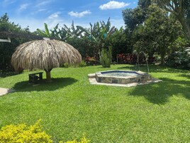 Un jardín con una estructura de techo de paja, un jacuzzi de piedra y abundante vegetación.