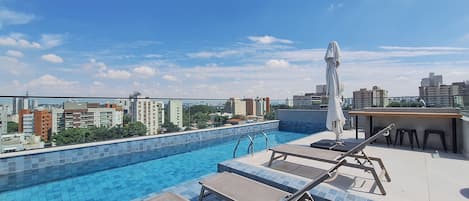 A rooftop pool with lounge chairs and a table, surrounded by a cityscape view.