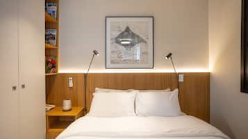 A neatly made bed with white linens, a wooden headboard, a bedside table with a lamp, and a shelf with books and a decorative item.