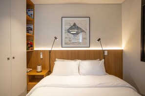 Ein ordentlich bezogenes Bett mit weißen Leinentüchern, ein hölzernes Kopfende, ein Nachttisch mit Lampe sowie ein Regal mit Büchern und einem dekorativen Gegenstand.