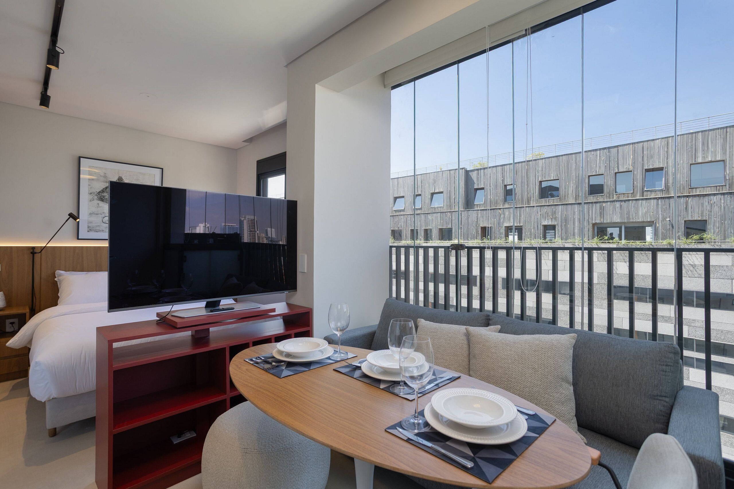 A modern hotel room with a bed, a TV, a dining area with a table set for two, and a balcony with a view of a building.