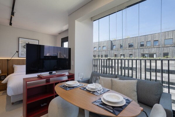 A modern hotel room with a bed, a TV, a dining area with a table set for two, and a balcony with a view of a building. - Vida Madalena (Sao Paulo)