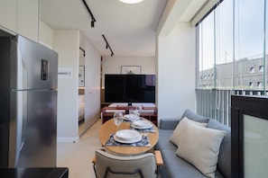 A modern kitchen with a built-in refrigerator, a dining area with a wooden table set for four, and a grey sofa with a cushion.