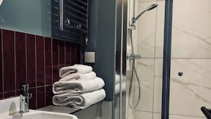 Une salle de bain équipée d’un lavabo, d’une pile de serviettes, d’une douche avec un pommeau de douche amovible et d’un luminaire fixé au mur.