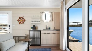 Una cocina compacta con lavamanos, estufa y armarios, un pequeño comedor con mesa y sofá, y un gran ventanal que ofrece vistas al agua y un puente.