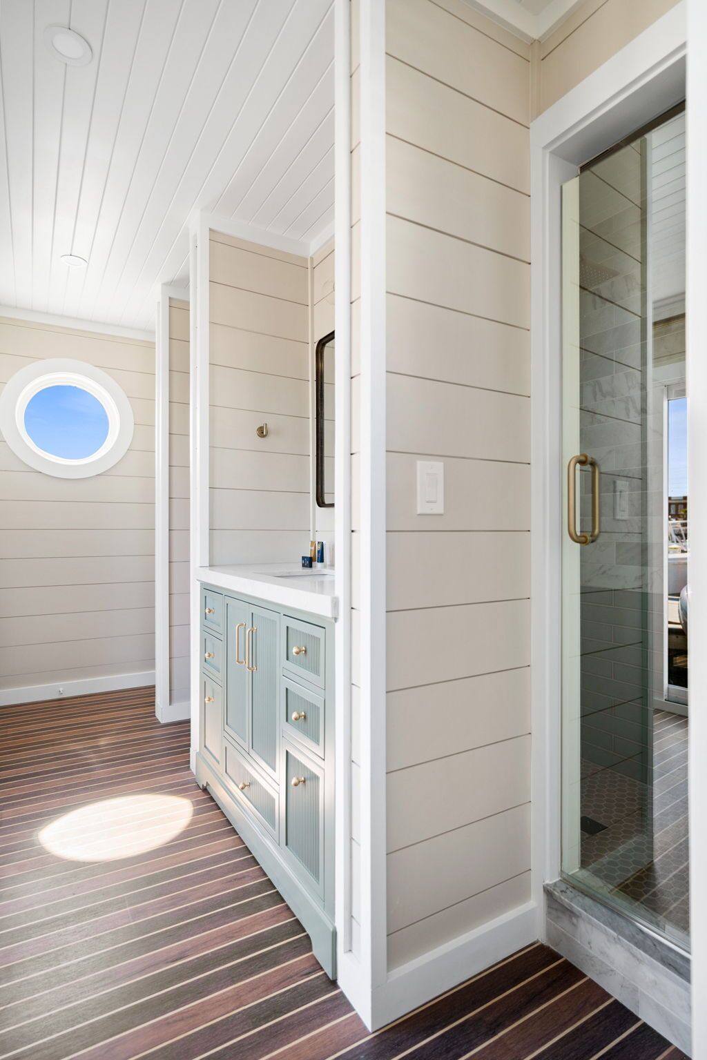 A room with a wooden floor, a round window, and a glass door leading to a shower area.