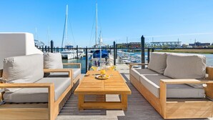 Una terraza de madera con mobiliario exterior y una mesa, con vistas a un puerto deportivo con barcos y un cielo azul despejado.