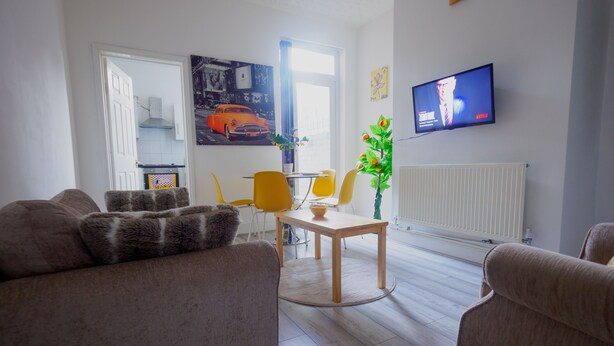 Een woonkamer met een bank, een eettafel met stoelen, een aan de muur bevestigde televisie en een raam met gordijnen.