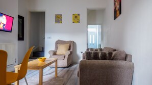 Een woonkamer met een bank, fauteuil en een tafel met een schaal. Aan de muur is een televisie bevestigd.