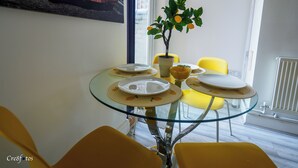 Een eetgedeelte met een glazen tafel, gele stoelen, een potplant en een schilderij van een auto aan de muur.