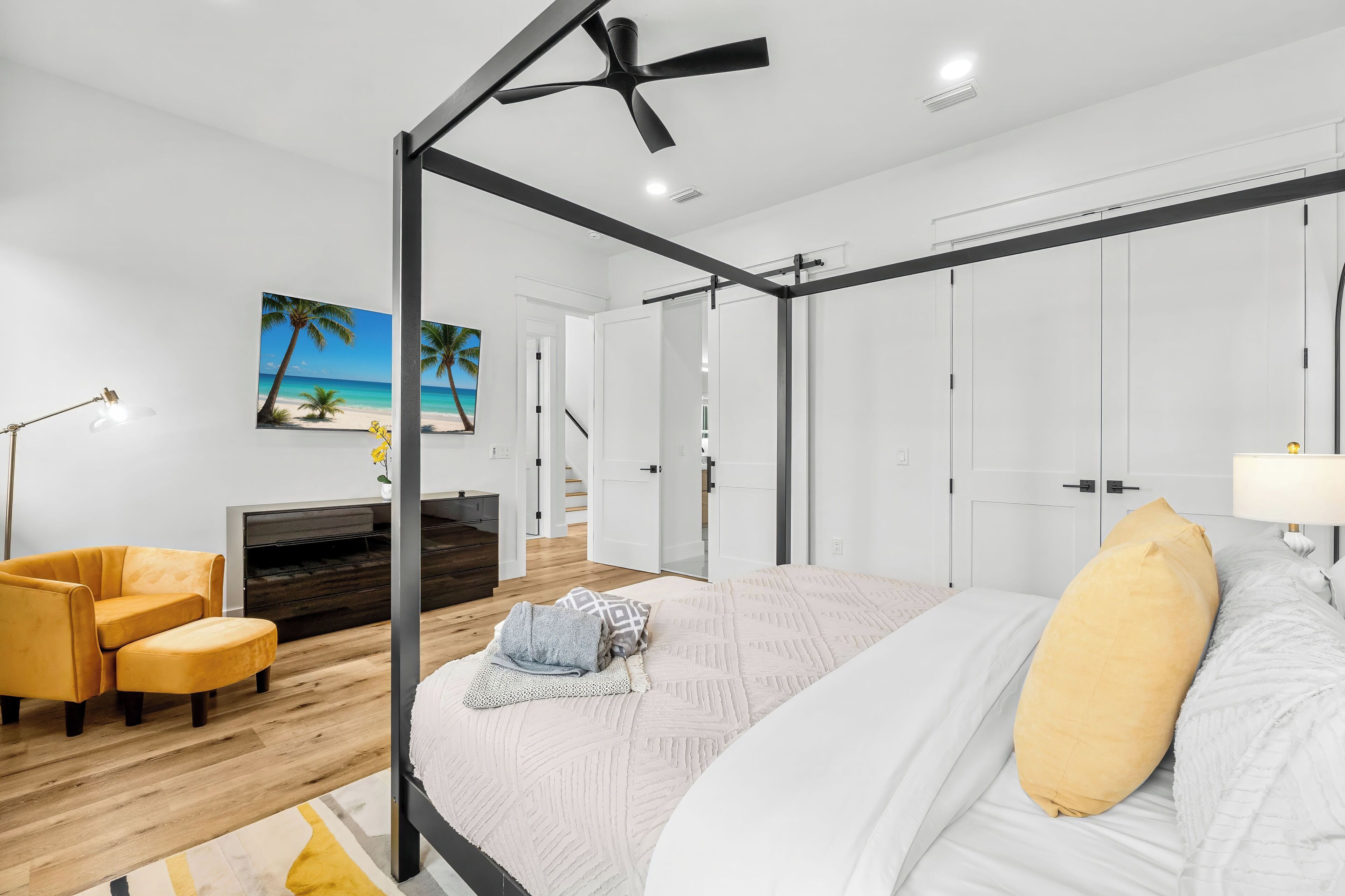 A modern bedroom with a four-poster bed, a wooden floor, a yellow armchair, and a painting of a beach scene.