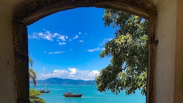 A view through an archway of a coastal area with boats, a pier, and people on the beach. - Amplo apartamento em Palmas (Governador Celso Ramos)