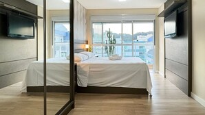 A modern hotel room with a large bed, a TV on the wall, and a view of buildings through large windows. - Amplo apartamento em Palmas (Governador Celso Ramos)