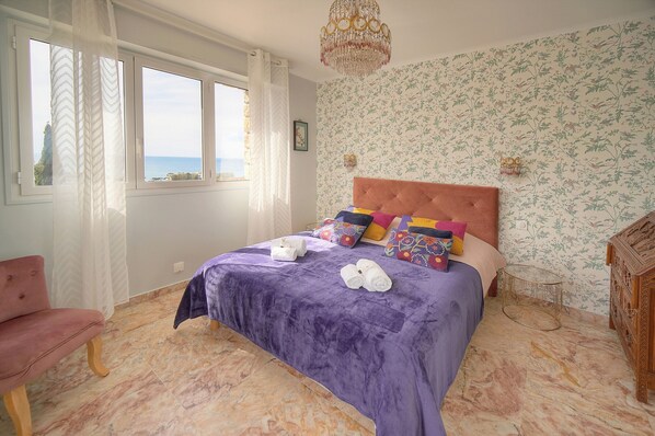 A bedroom with a bed, a chair, a chandelier, and a view of the sea.