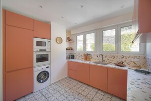 A compact kitchen with a washing machine, microwave, and oven. It features a window with a view of greenery outside.