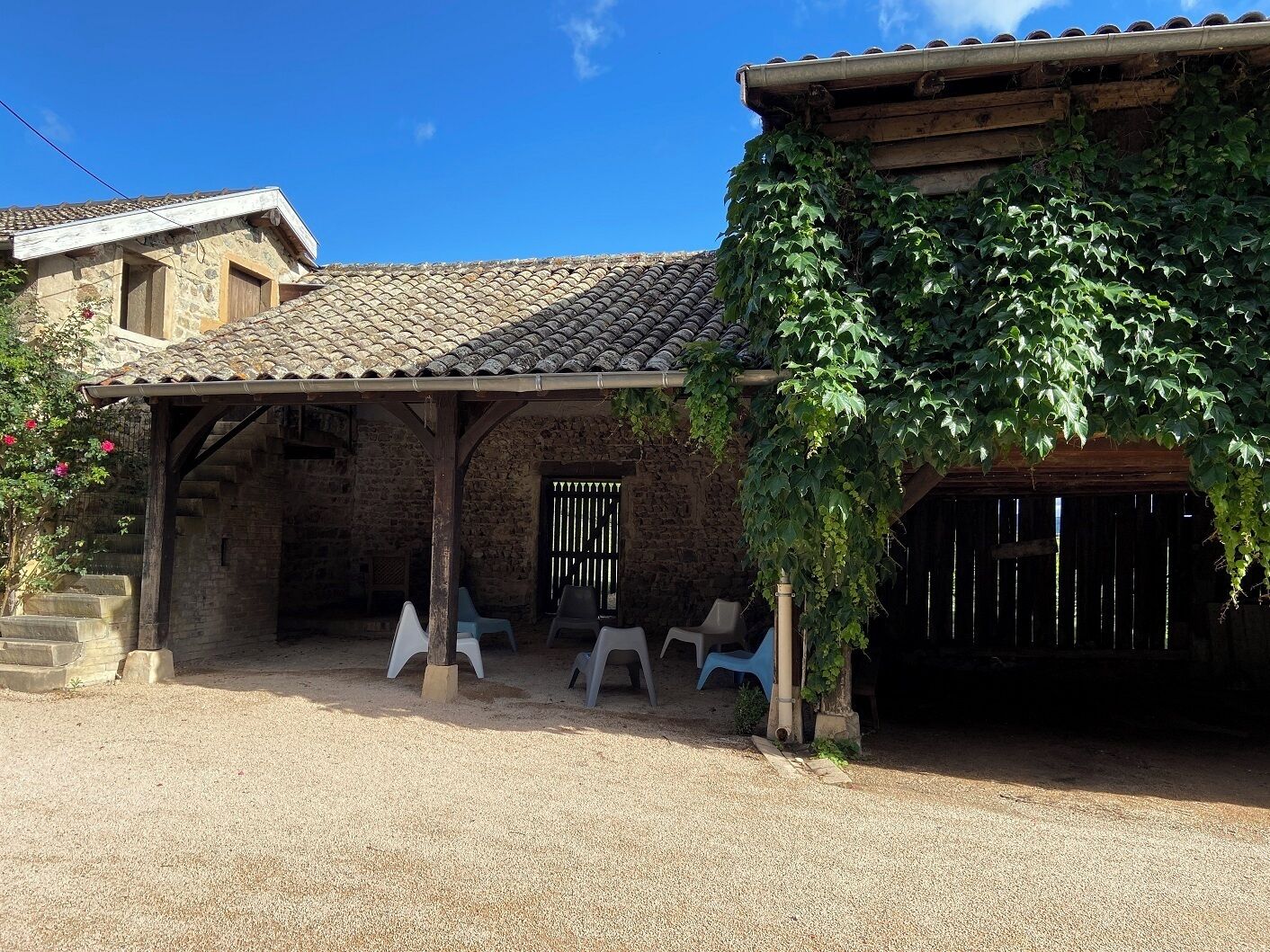 Edificio de piedra rústico con techo de tejas, un patio cubierto con sillas de plástico y suelo de grava.