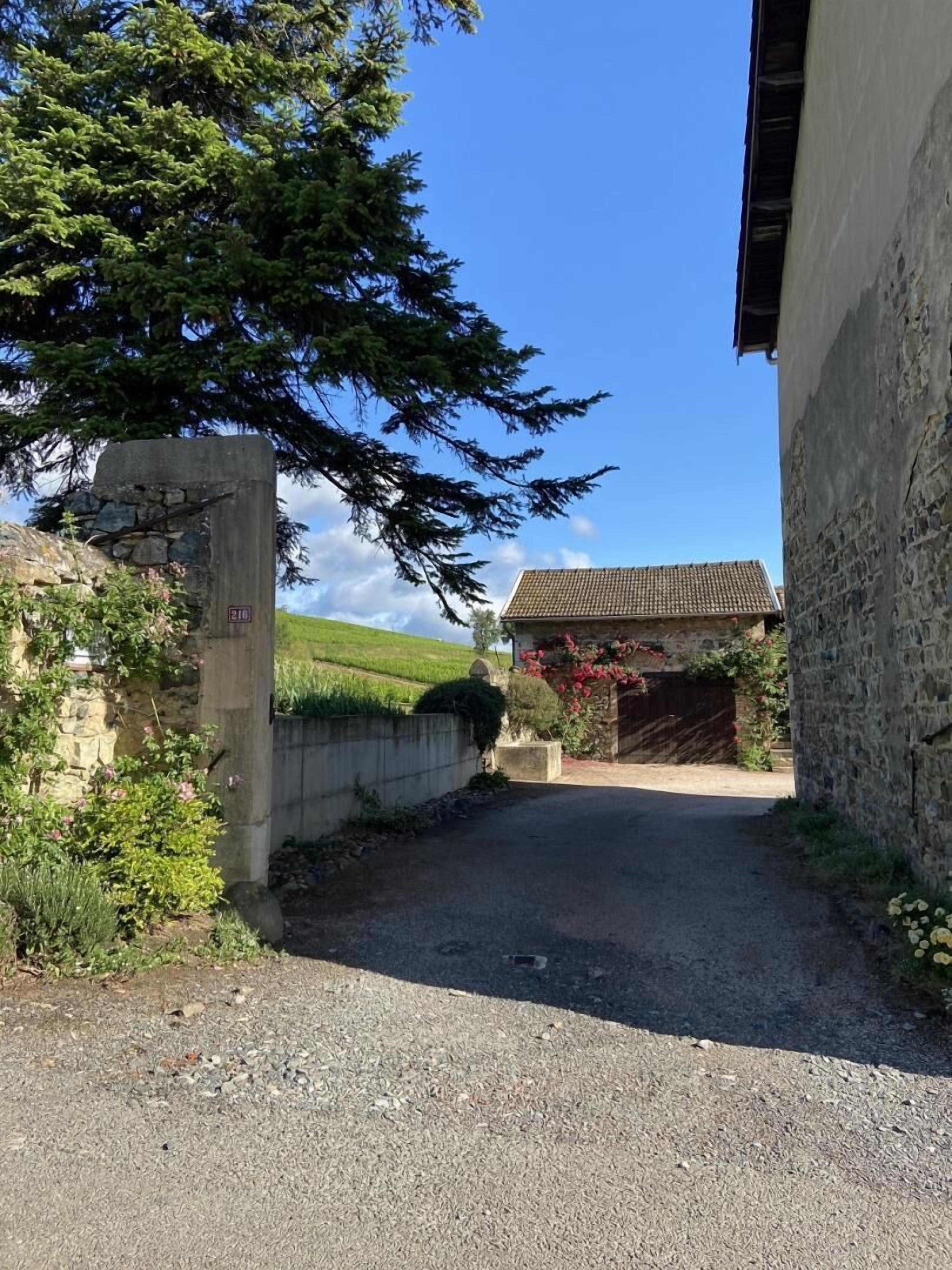 Un chemin de campagne mène à un bâtiment en pierre surmonté d’un toit de tuiles, entouré de verdure et sous un ciel dégagé.