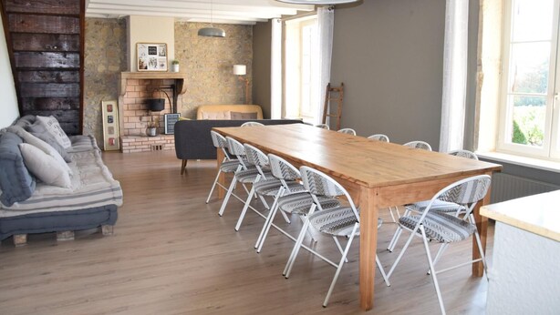 A spacious room with a wooden dining table surrounded by white chairs, a sofa with patterned cushions, a stone wall, and a large window providing natural light. - Gîte des Fournelles - SAINT-LAGER (SAINT-LAGER)