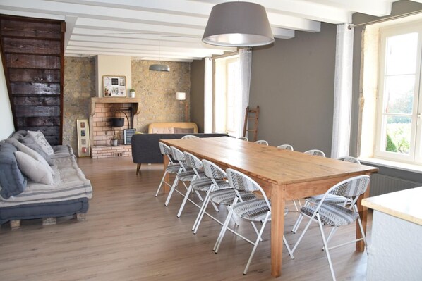 A spacious room with a wooden dining table surrounded by white chairs, a sofa with patterned cushions, a stone wall, and a large window providing natural light. - Gîte des Fournelles - SAINT-LAGER (SAINT-LAGER)