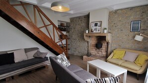 A cozy living room with a wooden staircase, a stone wall, a fireplace, and a white table. - Gîte des Fournelles - SAINT-LAGER (SAINT-LAGER)