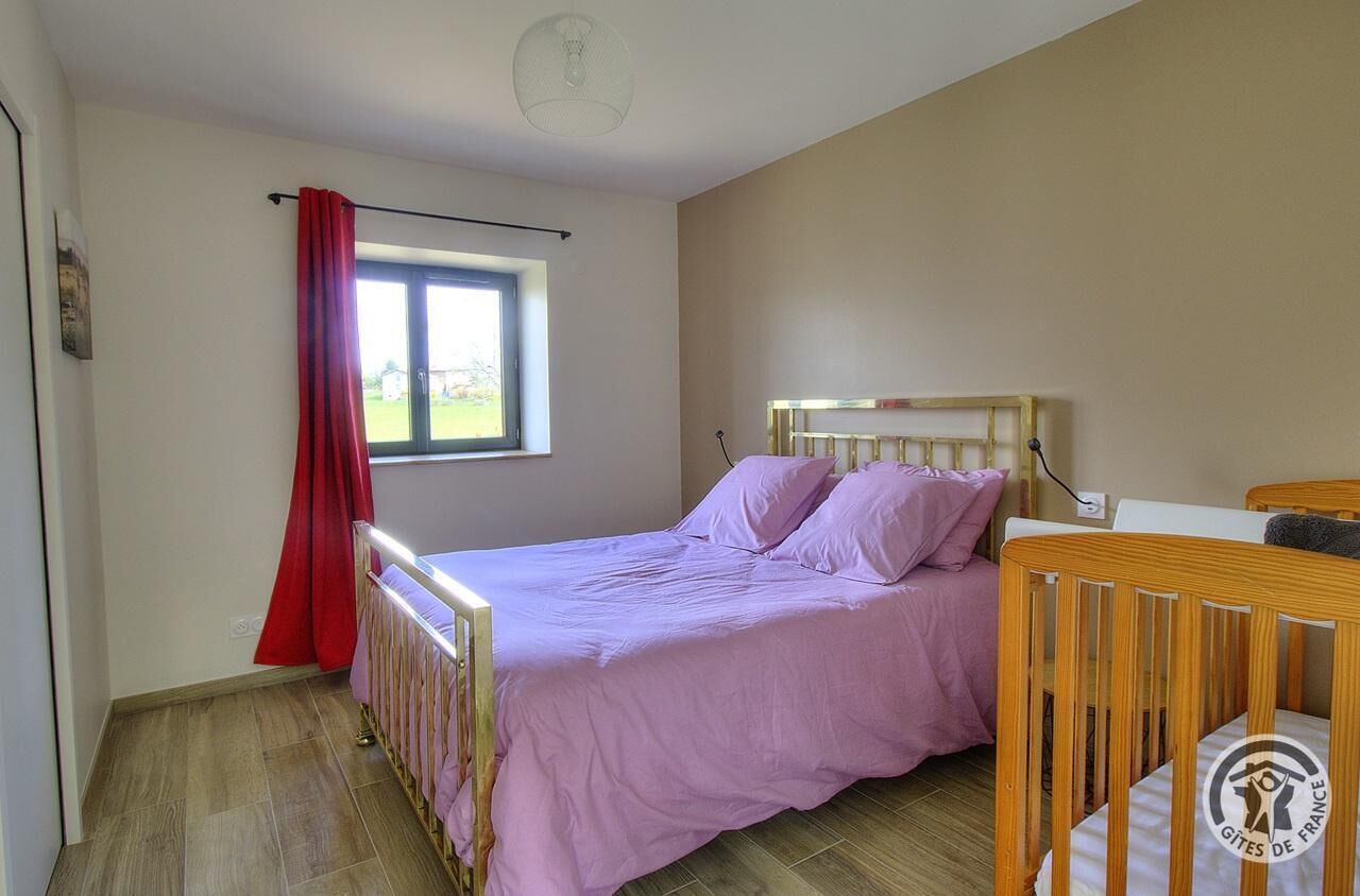 A bedroom with a bed, a crib, a window with a red curtain, and a light fixture.