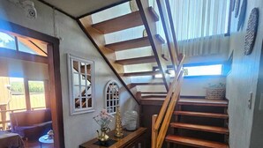 Una escalera de madera que conduce a un piso superior, una pequeña mesa con objetos decorativos y vistas al exterior.