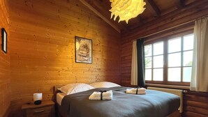 A wooden cabin bedroom with a bed, a nightstand, a lamp, and a window with curtains. - VACATION HOUSE VACATION APARTMENT EIFEL-ARDENNEN LOG CABIN VIEBIGHAUS (Buchet)