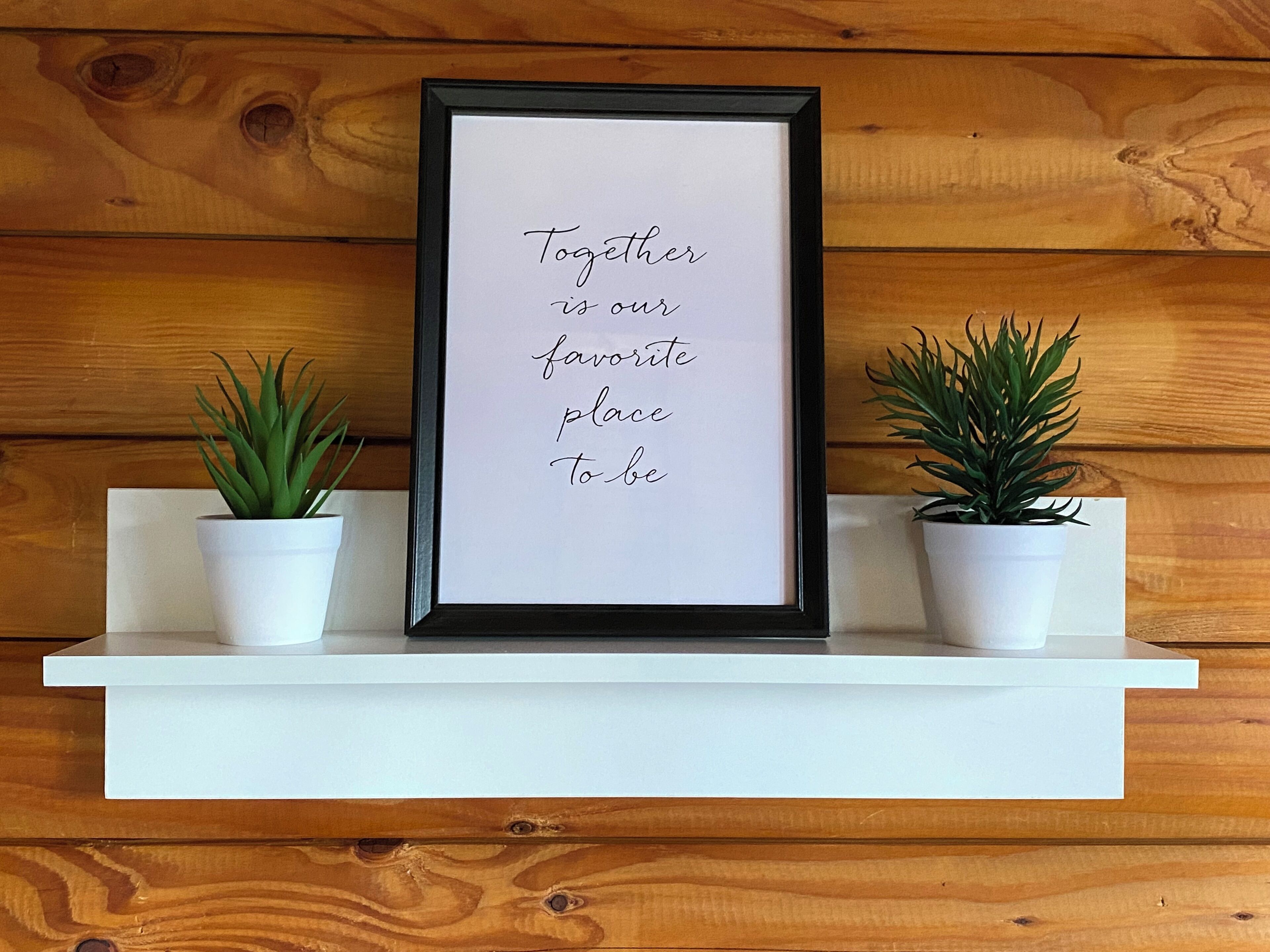 A framed quote on a white shelf against a wooden wall, flanked by two potted plants.