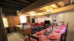 Uma sala de jantar com uma mesa comprida posta para várias pessoas, vigas de madeira no teto e uma cozinha com armários e pia.