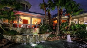 A luxurious house with a stone waterfall and a pool, surrounded by palm trees and a well-lit interior.