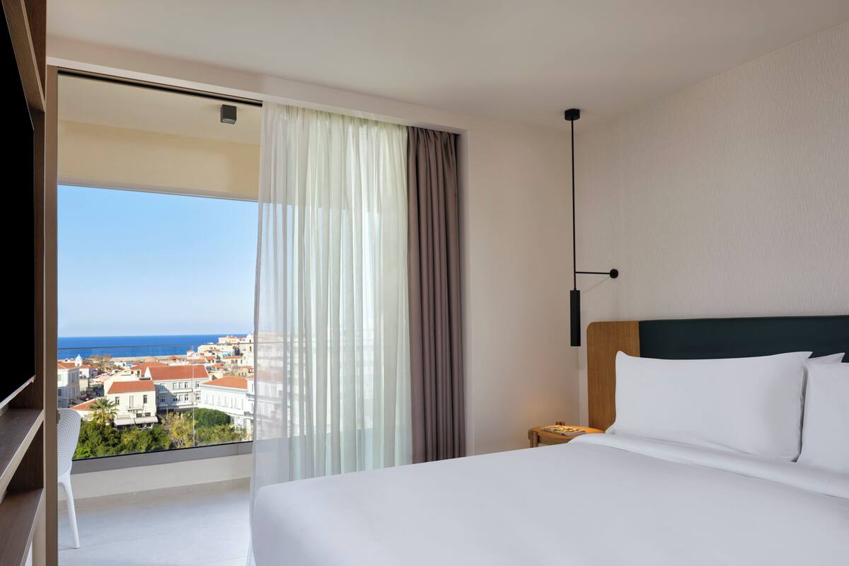 A hotel room with a large bed, a nightstand, a lamp, a view of the sea, and a balcony with a chair.