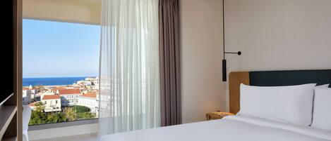 A hotel room with a large bed, a nightstand, a lamp, a view of the sea, and a balcony with a chair.